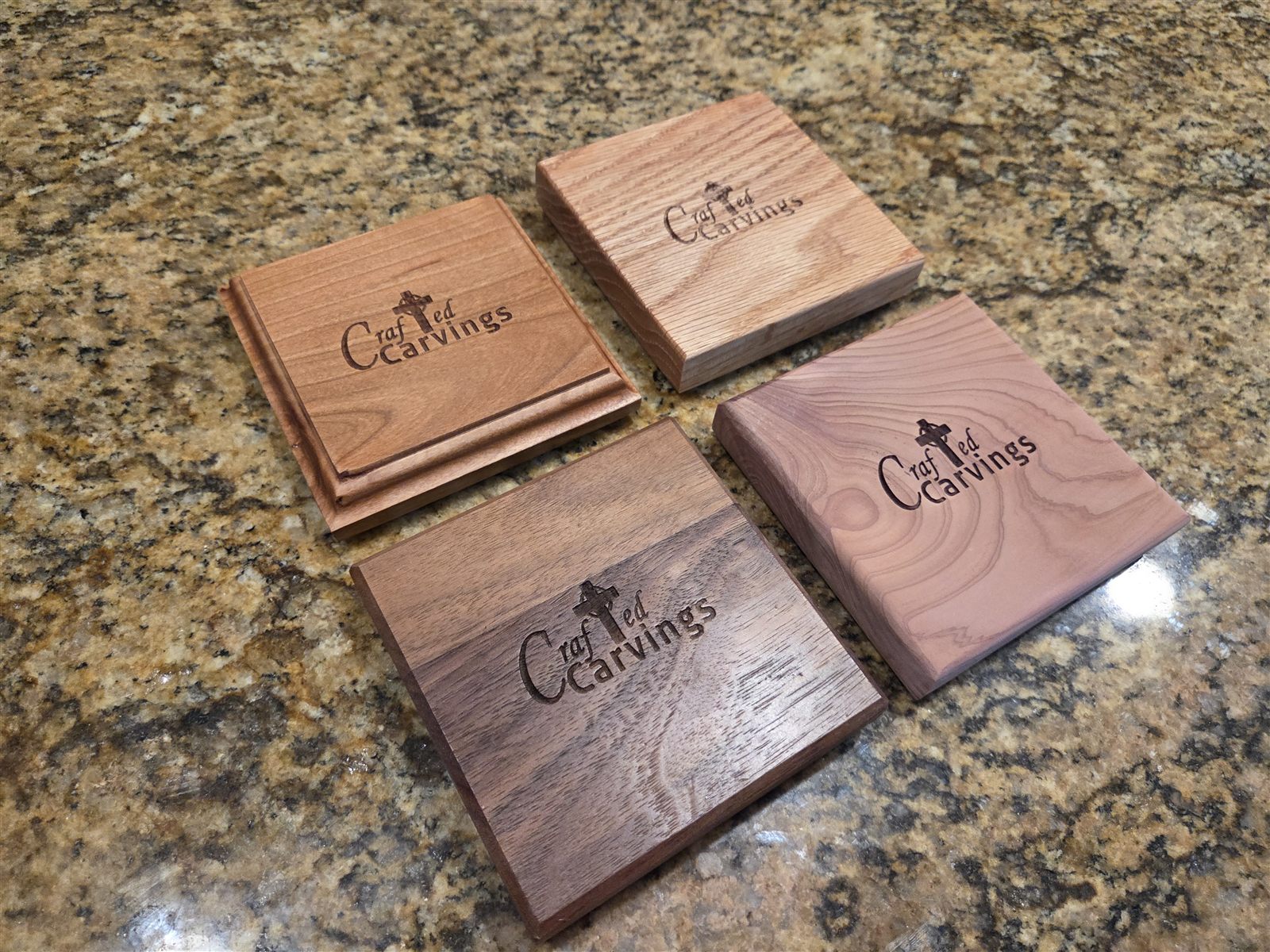Four square wooden Keepers in four different woods on a granite counter, each engraved with the Crafted Carvings logo